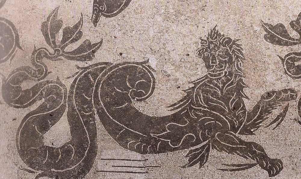 A mosaic of a lion-sea creature. Its upper body is lion-like, but its lower half is a long fish tail, coiled around itself.